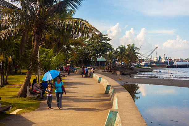 Davao, Philippines Waterfront Davao, Philippines - April 26, 2012: People sit in a public part along the waterfront under a coconut palms while a woman and child walk along the waterfront esplanade. davao city stock pictures, royalty-free photos & images