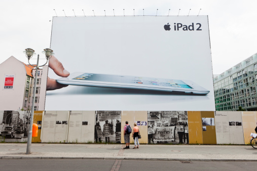 Berlin, Germany - May 11: A large advertisement for the Apple iPad 2 on Friedrichstrasse near Checkpoint Charlie and photo exhibition in Berlin, Germany. Couple reading documentary text about the Wall for period 1980-89. Apple ad is behind the fence.