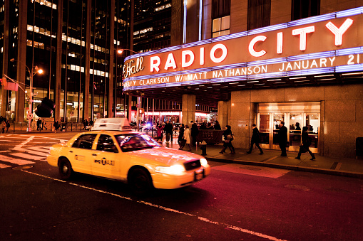 New York, United States - January 11, 2012: People and taxis passing by the Radio City Music Hall at night in New York. This famous entertainment venue is located at the Rockefeller Center and is home of different entertainments events from music to sports.