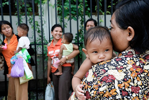 Phnom Penh, Cambodia - June 19, 2007: A Cambodian woman holds her baby as they wait in a long line outside a clinic for free child vaccinations. Other mothers and children waiting in line are in the background.