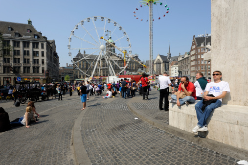 Amsterdam, the Netherlands - April 22, 2011: View from the National Monument (to the right) at a carnival on Dam Square in Amsterdam. Dam Square lies in the historical center of Amsterdam and is one of the city's most well known places. It's a meeting place and a location for many events. The National Monument is a Second World War monument. In the picture, people are sitting on the monument, relaxing in the sun.
