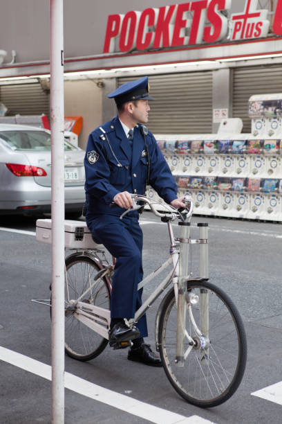 Madrid, Spain, Europe, September 11, 2025: Daytime urban scene in central Madrid featuring a Spanish police officer standing next to a marked BMW motorcycle parked on a sidewalk. The officer, dressed in a dark blue uniform and cap, appears engaged with a mobile device while overseeing the area. The motorcycle displays high-visibility markings and emergency contact numbers, positioned near a tree-lined street with stone buildings and barred windows.