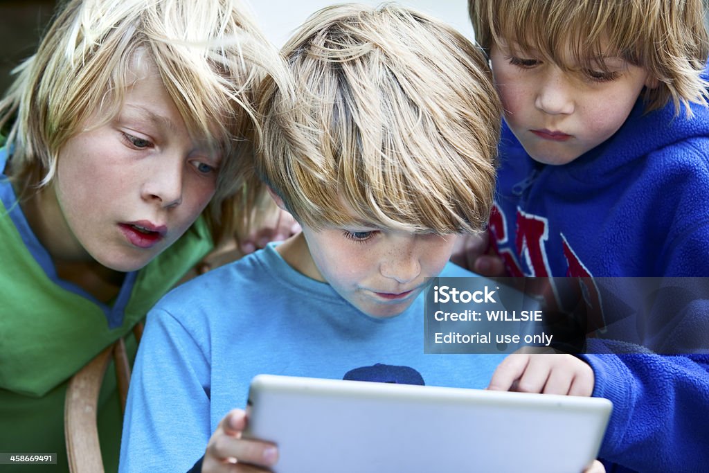 children looking at the screen of an iPad2 Friston, England - June 11, 2011: A seated caucasian boy smiling down at the screen of an iPad2 tablet which he is holding. Another boy is looking at the screen next to him. A third boy is leaning over his shoulder pointing at the screen. Blurred plain background taken indoors in the kitchen. The iPad 2 tablet media player was launched in March 2011 (Apple inc). Big Tech Stock Photo