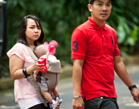 Singapore, Singapore - January 22, 2012: Chinese mother keeping her baby cool with battery driven fan. Young couple walking in tropical heat. Mother carries baby in baby carrier and tries to keep him/her cool with a battery driven fan.