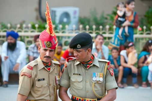 Wagah, Punjab, India - July 10th, 2011: Two Indian soldiers, one with red turban, talking before the ceremony called the Lowering of the Flags on the India-Pakistan border. Behind them Indian audience(woman with child standing, Sikh on the left sitting) can be seen.