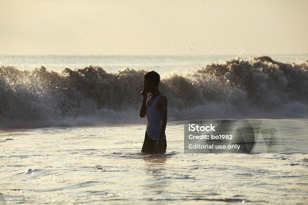 young man standing in water on the beach Bali,Indonesia-April 30,2010: one young man standing in the water on the beach while the wave coming at the background. Adult Stock Photo