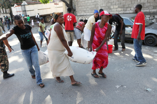 Port-Au-Prince, Haiti - February 7th, 2010 : Three women help each other carry two sacks of rice which are being rationed by the USAid - an American relief aid organisation. In the background other families struggle with their heavy rice sacks as a UN soldier overlooks the rationing.