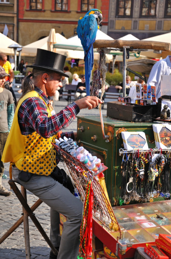 Warsaw, Poland - October 1st, 2011: Senior organ-grinder in yellow jacket and black top hat, playing on his hurdy-gurdy full of souvenirs from Warsaw. Blue parrot sitting on a stick above the hurdy-gurdy. Restaurant umbrellas and rchitecture of the Old Town in the background.