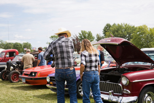 Fosston, USA - August 27, 2011:  A man and a woman wearing almost-matching plaid shirts and blue denim jeans (his Levis, hers Wranglers), are walking and viewing a row of collector\\'s cars at a classic car show.  Additional people and vehicles are in the background as well as reflected in the chrome bumper of the front car.