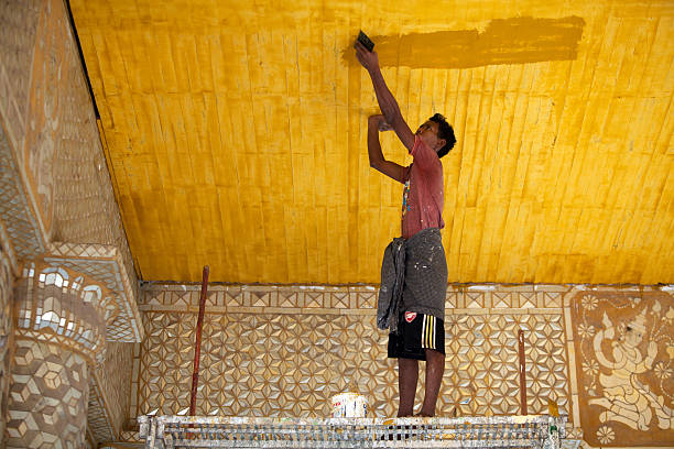 Painting a temple, Myanmar Amarapura, Myanmar - February 19, 2011: A young man stands on a temporarily built framework and colours the ceiling of a temple a bright yellow gold. Amarapura, the former capital of Myanmar, means City of Immortality in Pali. It was founded after the court astrologers adviced the King to move the capital of the country to Amarapura after which the entire population of Inwa was asked to pack their belingings and migrate to the new land. Today the city is the center of weaving activities and is known to produce some of the finest festive clothing in Myanmar. Amarapura stock pictures, royalty-free photos & images