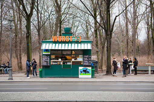 Berlin, Germany - January 5, 2012: Kiosk for sausages in Berlin, Germany. Located between Brandenburg Gate and Reichstag (Parliament buildings) at the Tiergarten park this kiosk offers the famous Berlin Currywurst a sausage served with a spicy tomato sauce with curry and sometimes french fries. In addition the kiosk offers drinks and other variations of sausages. Several customers are enjoying their food around the kiosk.  This image shot in winter.
