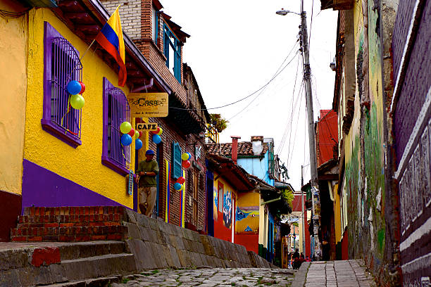 Bogotá Colombia - The Narrow, Colorful, Cobblestoned Calle del Embudo In The Historic La Candelaria District. Spanish Colonial Architecture. Bogota, Colombia - July 20, 2013: Cobbled street in La Candelaria known as Calle del Embudo, in the capital city, Bogota, Colombia in South America; homes and restaurants are painted in the bright colours of Latin America. It is national day and the Colombian flag and balloons can be seen on a window and walls. Very few people are on the streets at this time; it will come alive in the evening. calle del embudo stock pictures, royalty-free photos & images
