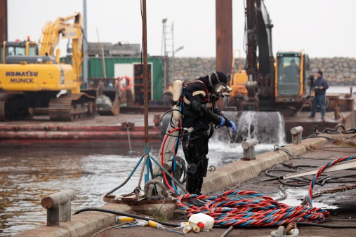 Marstal, Denmark - November 23, 2011: Commercial diver ready to enter water from pier. In the background excavators are used to deepen the harbor.