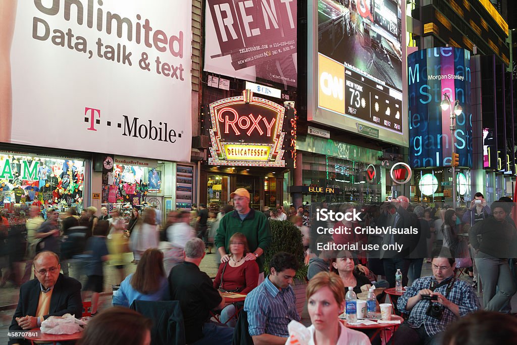 Times Square crowds and advertising billboards New York, NY, USA - October 8, 2011: Crowds of tourists walk south down Broadway into Times Square after dark to see the lights, go to the theater, and eat. Huge billboards on the buildings along Seventh Avenue and Broadway advertise theater shows, products and services. In the background there are ads for T-Mobile cellular service, the Roxy Delicatessen, the show Rent, CNN, and Morgan Stanley financial services. Visitors and New Yorkers are enjoying a sit down at the tables and chairs provided as a rest station in this photo. The people are blurred in this long exposure. Arts Culture and Entertainment Stock Photo