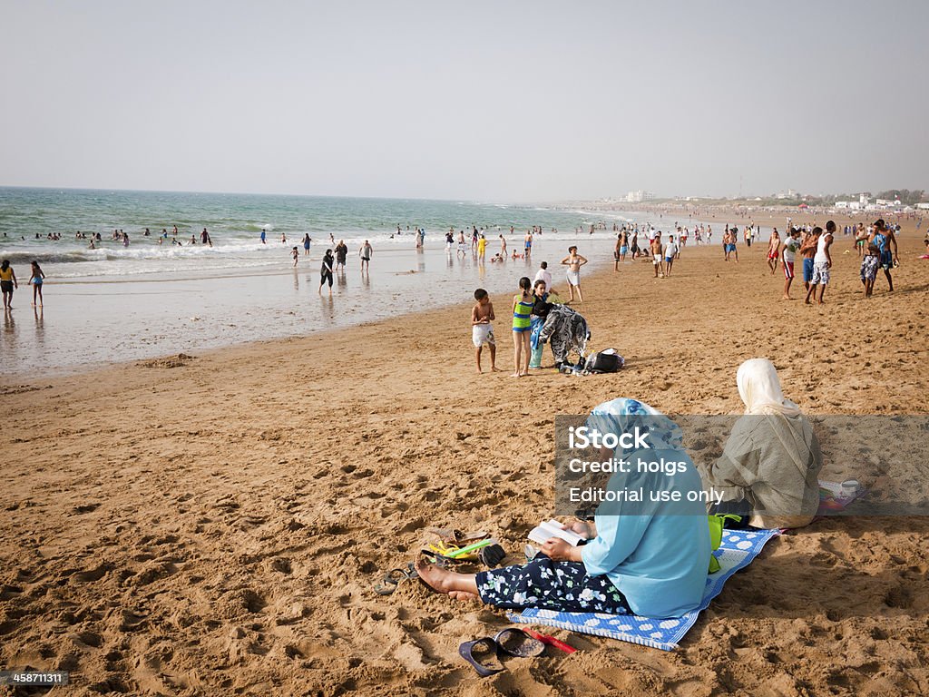 Asliah, Morocco - woman wearing headscarves sit on the beach Asliah, Morocco - July 9, 2010: Moroccan woman wearing headscarves sit on the beach in the late afternoon with other locals playing in the background Asilah Stock Photo