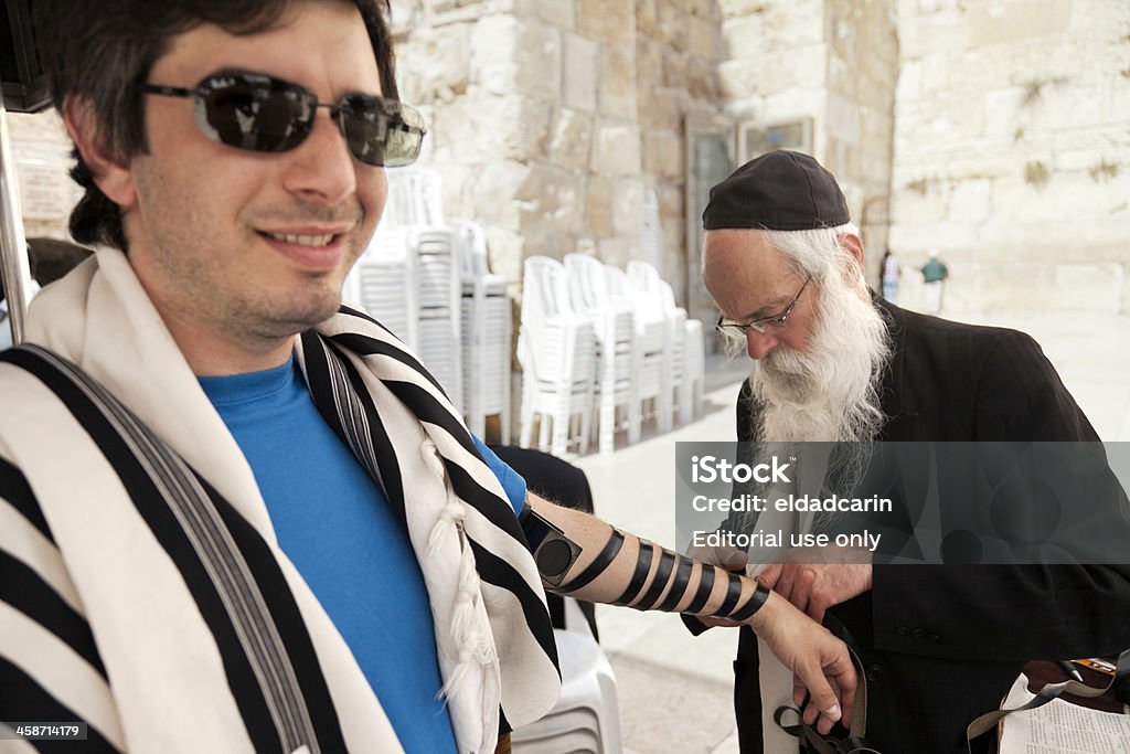 Tourist Wearing Phylacteries "Jerusalem, Israel - May 9th, 2012: An adult Caucasian man wearing a praying shawl , while being helped by a Rabbi with putting on the Phylacteries (Teffilin, Tefilin, Tfilin). This man is a Jewish tourist visiting the holy Western Wall (aka Wailing Wall) at the old city of Jerusalem. The Rabbi is practicing his heavenly duties by helping people engage in the phylacteries ceremony." Active Seniors Stock Photo