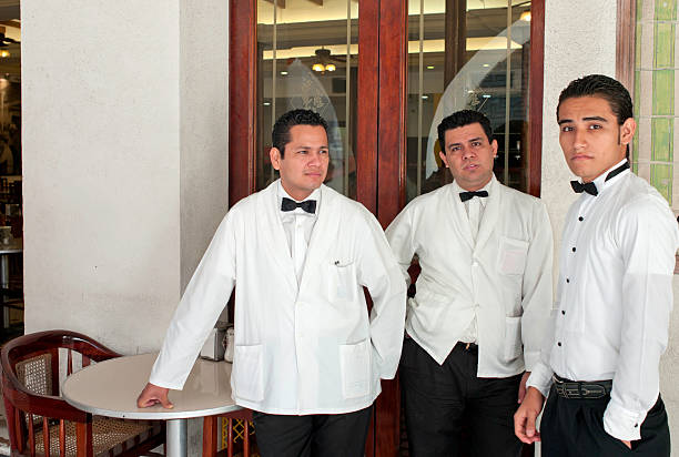 Holds empty plate. Black waiter in formal clothes is at his work outdoors at sunny daytime.
