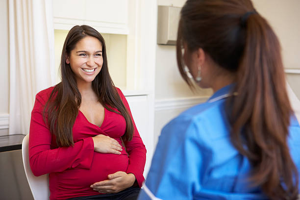 A pregnant woman holding her stomach in front of a nurse Pregnant Woman Meeting With Nurse In Clinic to discuss pregnancy woman talking to doctor stock pictures, royalty-free photos & images