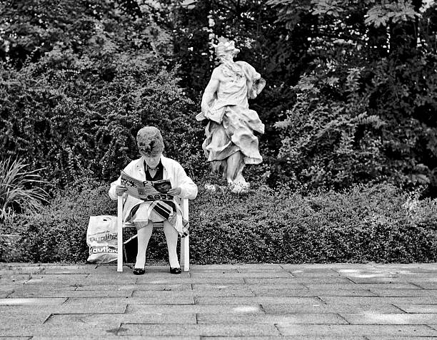 Augsburg, Germany - April 11, 1988: A middle aged lady sitting in a public park reading a magazine. Close by to the right in the background a stone statue of a male classical mythological figure, head inclining to the left, appears to be looking over her shoulder.