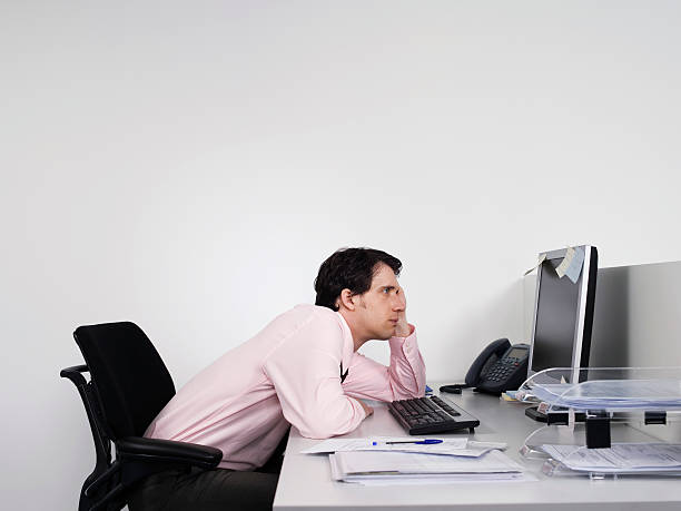 Bored Male Office Worker At Desk Side view of a bored male office worker looking at notes on computer monitor at desk bad posture stock pictures, royalty-free photos & images