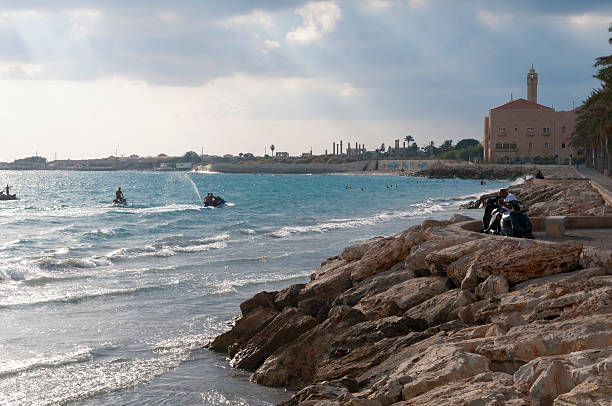 Tyre, Lebanon - September 1, 2010: Late in the afternoon, people enjoy the seaside in Tyre. People are using jet skis, swimming, and sitting on a bench. In the distance (center) are ancient columns at the Al-Mina Archaeological Site.