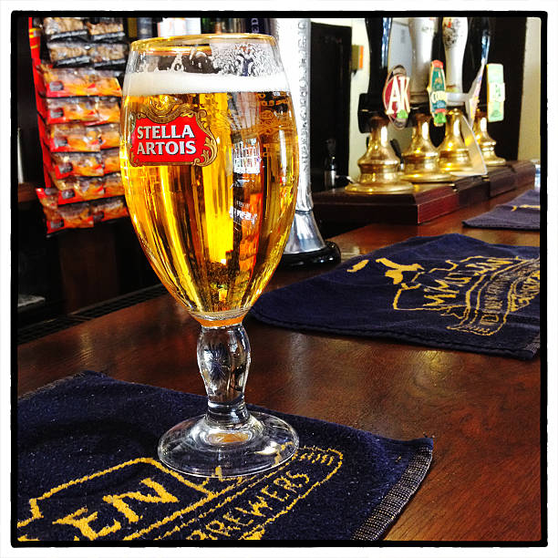 Stella Artois Beer on an English Pub Counter "Aston, England - September 6th, 2012 : A pint of Stella Artois beer on a traditional English pub counter. Beer pumps and packets of traditional pork scratchings are shown in the background. There are McMullen & Sons drip towels on the bar counter. McMullen have been brewing beer in the Hertfordshire area since 1891 and operate a large number of English pubs." pint of stella stock pictures, royalty-free photos & images