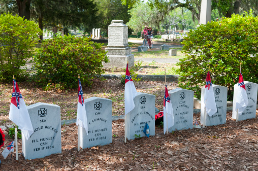 Charleston, South Carolina, USA - May 10, 2012: Two women (background) bike through historic Magnolia Cemetery on a summer day. In the foreground are the graves of Confederate Navy sailors who perished in 1864 aboard the H. L. Hunley, a submarine that made a mark in the history of naval warfare. It was the first combat submarine to sink an enemy warship. The Confederacy lost 21 crewmen in three sinkings of the Hunley during her short career.