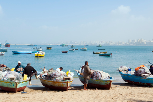 Alexandria, Egypt - February 28, 2011: Egyptian fisherman and their boats on the beach in Alexandria Harbor. Among the buildings in the distance is the famous Alexandria Library.