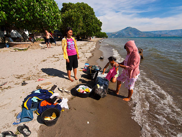 On lake "Samosir, Indonesia - June 9, 2009: Group of women from island Samosir is washing their family clothes in the waters of lake Toba on the beach. In background are green trees and blue sky." danau toba lake stock pictures, royalty-free photos & images