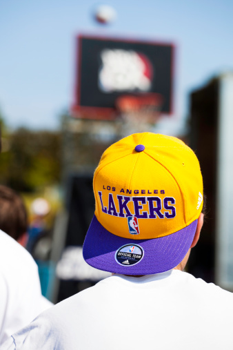 Halifax, Nova Scotia, Canada - September 8, 2012: A young Los Angeles Laker fan watches a game being played outdoors.  Los Angeles Lakers hat includes NBA logo and sticker on brim of hat.  Basketball hoop and backboard visible in background as well as a basketball that was shot towards basket.