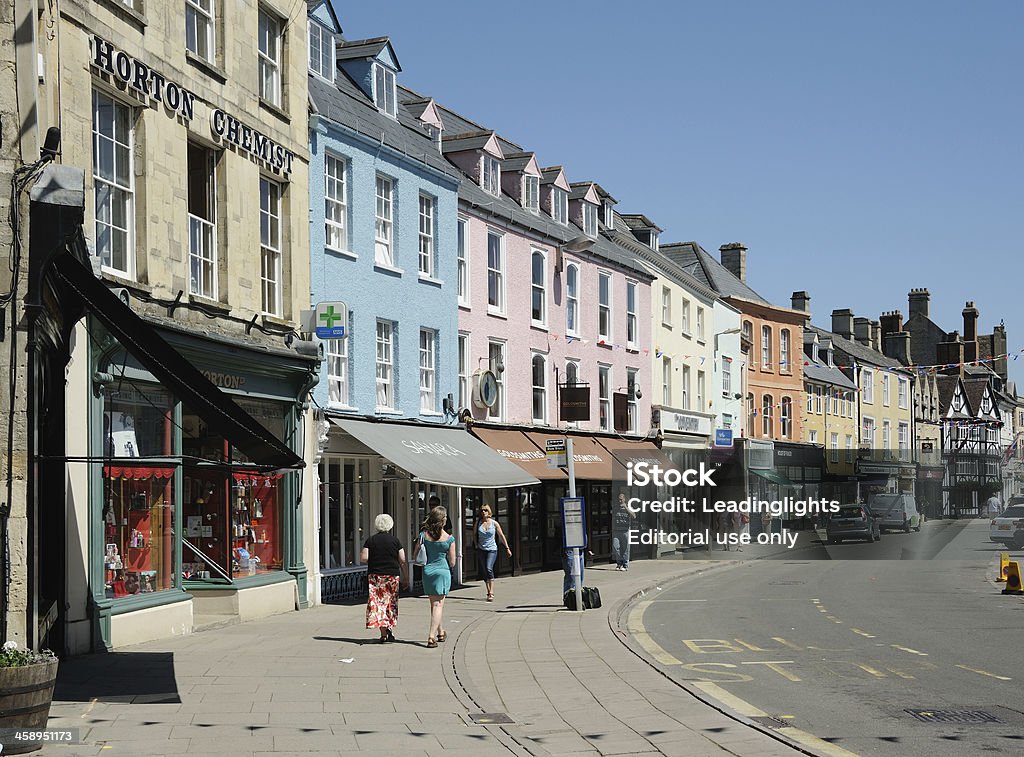 Cirencester Market Place "Cirencester, United Kingdom - May 26, 2012: North side of the Market Place, Cirencester, Gloucestershire on a sunny day in May. Unidentified people pass by pastel-coloured shops including a chemist, clothes shop, jeweller, building society and department store. Half-timbered Fleece Inn in the distance, bus stop in the foreground.." Cirencester Stock Photo