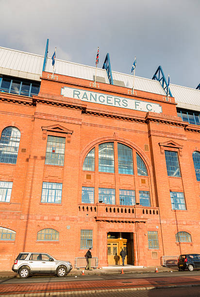 Ibrox Stadium, Glasgow "Glasgow, UK - December 7, 2012: Evening light on the Rangers F.C. sign over the entrance to the Bill Struth Main Stand at Ibrox Stadium, Glasgow, the home ground of Rangers Football Club. The main stand was built in 1928 with an impressive red brick facade. A man stands outside the entrance talking on a phone, whilst another man enters the building." ibrox stock pictures, royalty-free photos & images