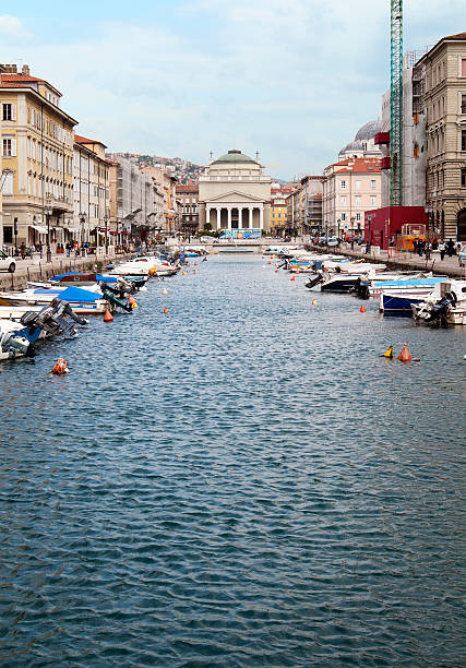 Trieste, Italy - May 6, 2010: Boats moored and pedestrians walk alongside the Grand Canal, with the Sant'Antonio Nuovo in the background.