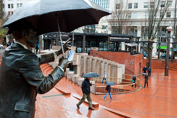 Portland, Oregon, USA - April 3, 2012: On a rainy day in downtown Portland, pedestrians walk through Pioneer Courthouse Square. A Starbucks is in the background. In the foreground is one of the most recognized pieces of public art in the city, a bronze statue created by Seward Johnson. Commonly referred to as Umbrella Man, it was given to the city in 1963.