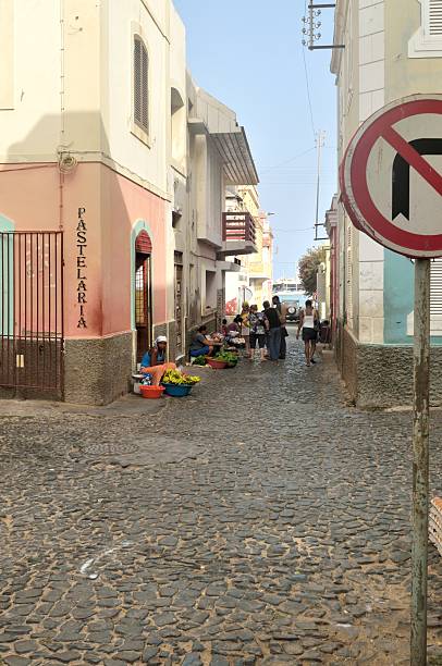 Mindelo, Cape Verde - May 23, 2012: Mindelo women operate farmers markets to sell their produce in a cobblestone paved alleyway with both locals and tourist as customers. The group of people standing on the left are passengers from a cruise ship docked for a one day excursion.