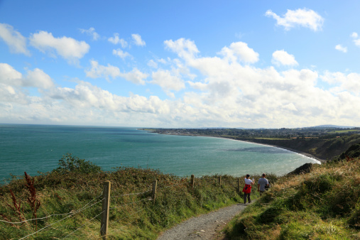 Greystones, Ireland - September 9, 2012: On a bright September day, a couple walk the scenic coastal path between the towns of Greystones and Bray in County Wicklow, Ireland.  The view is south towards the town of Greystones. The couple are carrying wet weather clothing and a plastic bottle of water.