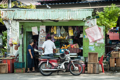 Penang, Malaysia - August 7, 2011: Chinese Malaysian Women selling convenience items from a corrugated shop on the streets of Penang. Local man buying from store and his parked motorcycle.