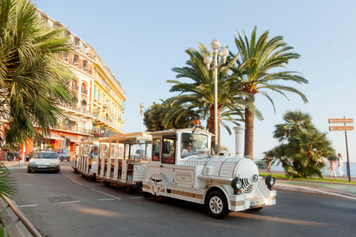 Nice, France - July 01, 2011 : Nice street live at sunset - white fun trackless train for sightseeing in front, one car at the background and pedestrians walking on Promende des Anglais