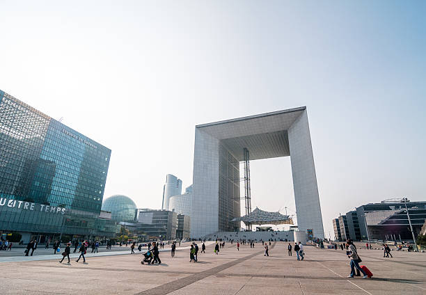 Wide view of people walking through the plaza in La Defense business district, with the Grande Arche and modern office buildings in the background on a cloudy day, Paris, France June 01, 2025