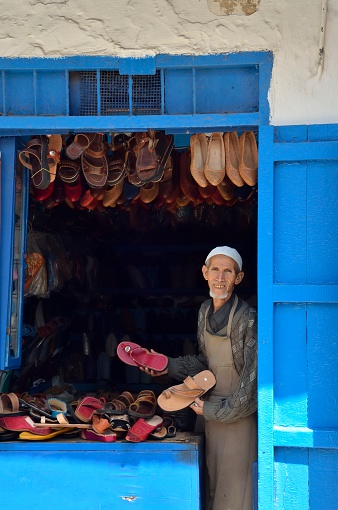 Rabat, Morocco - May 28, 2012: A senior man, a cobbler or shoe vendor, offers his goods to tourist from a darkened alcove in the medieval alleyways known as the Kasbah des Oudaias in Rabat, Morocco.