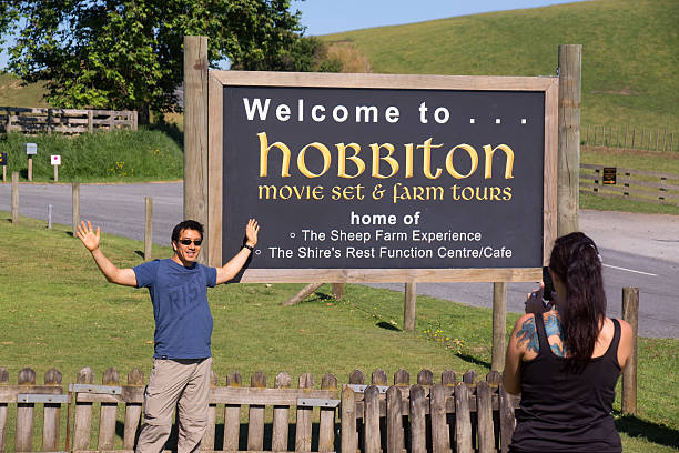 Hobbiton - Welcome Sign "Matamata, New Zealand - January 7, 2013: A visitor poses for a photo before a welcoming sign to Hobbiton. Hobbiton features an actual movie set of The Shire, used in the filming of the Lord of the Rings trilogy and The Hobbit." matamata new zealand stock pictures, royalty-free photos & images