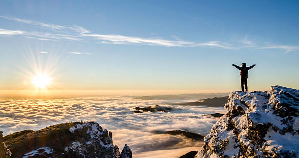 success in a majestic sunrise hiker enjoys success on top of a cliff above the sea of clouds. in front of him there is the rising sun spreading its warm beams over the land arms outstretched photos stock pictures, royalty-free photos & images