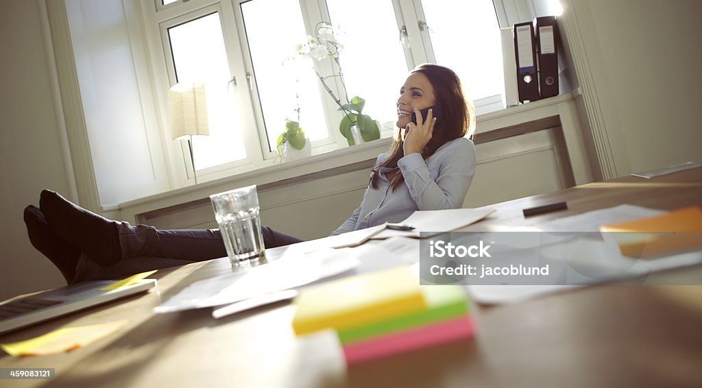 Beautiful business woman talking on cell phone Beautiful young businesswoman sitting at her desk with legs on table talking on mobile phone smiling. Caucasian young female working in home office. Feet Up Stock Photo