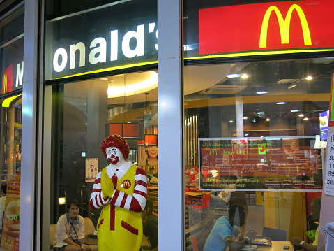 Bangkok, Thailand – December 16, 2013: A  View of Ronal McDonal in Bangkok, Thailand.  Ronald McDonald poses with the traditional Thai greeting called a 'wai' in front of McDonal’s. There are some advertisements of McDonal’s on the windows. Customers are inside  the restaurant.