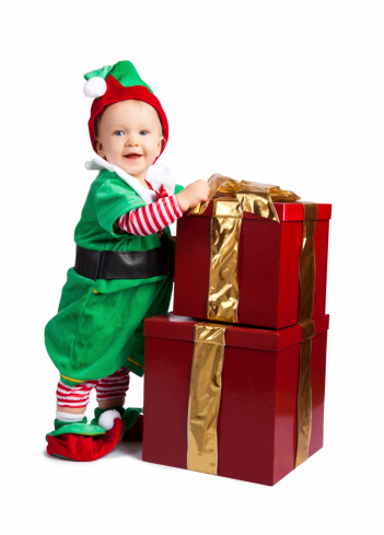 An adorable blue eyed baby dressed as an elf standing next to two large Christmas presents.  Isolated on a white background.