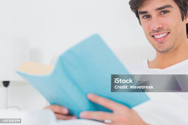Alegre Hombre Sentado En La Cama Libro De Lectura Mirando A La Cámara Foto de stock y más banco de imágenes de 20 a 29 años