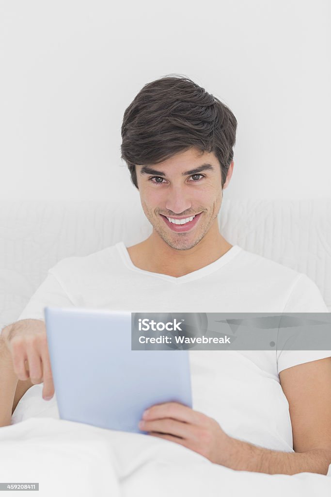 Feliz homem sentado na cama, usando tablet olhando para a câmera - Foto de stock de 20 Anos royalty-free