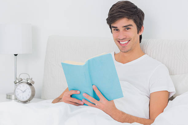istock Young man sitting in bed holding book looking at camera 459208453