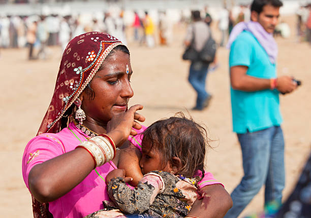 Pushkar, India- November 15, 2010: Indian woman nursing child. The place is Pushkar were the yearly camel market is held and draws lots of people, In the background people are standing an walking by. It is hot, Note the sweatdrops on the mothers forehead.