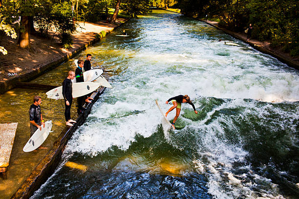 Munich, Germany - August 26, 2011: Young woman surfing with other people watching at the Eisbach river in Munich Germany. The Eisbach river is a small but fast and cold stream of water tributary of the Isar river that flows through the Englischer Garten park where surfers gather to practise their sport specially at this spot were the river forms a standing wave
